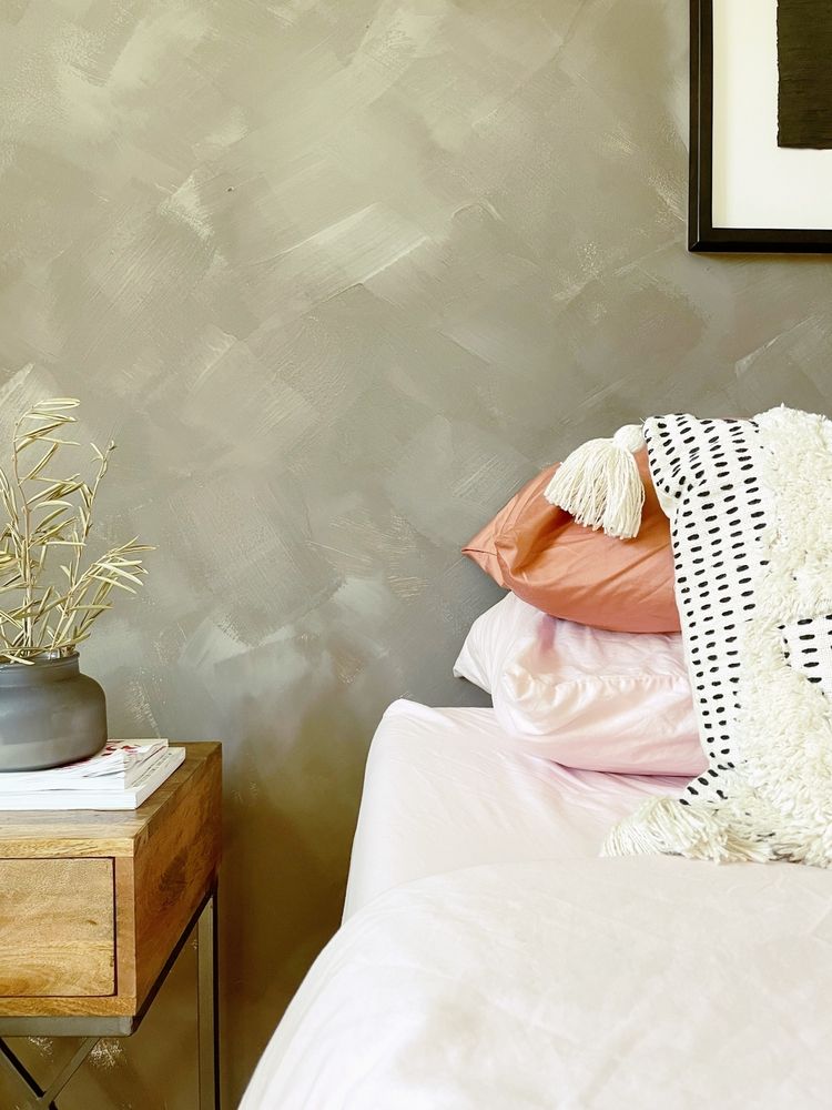 Bedroom with textured gray wall, nightstand, and bed with pink and peach pillows, cream throw.