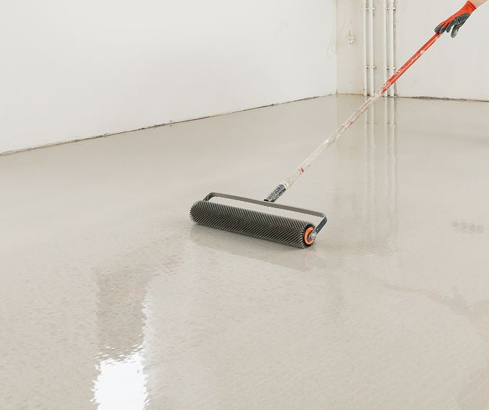 Person rolling a textured roller across a light gray epoxy floor in a white-walled room.