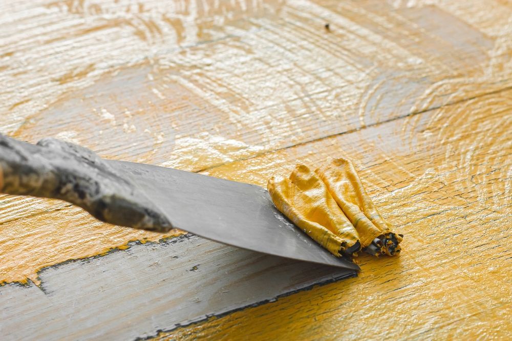 A metal putty knife scraping yellow substance off wooden planks.