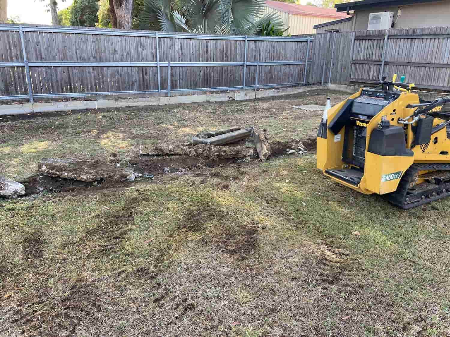 A yellow tractor is digging a hole in the grass in a backyard.