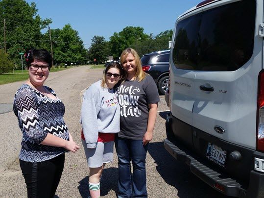 Three women are standing next to a white van in a parking lot.
