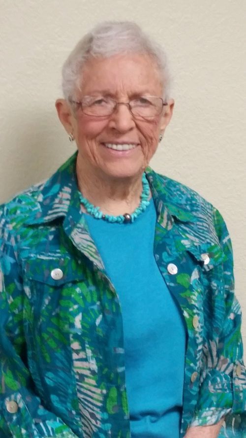 An elderly woman wearing glasses and a blue shirt is smiling for the camera.