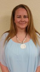A woman wearing a light blue top and a seashell necklace is smiling.