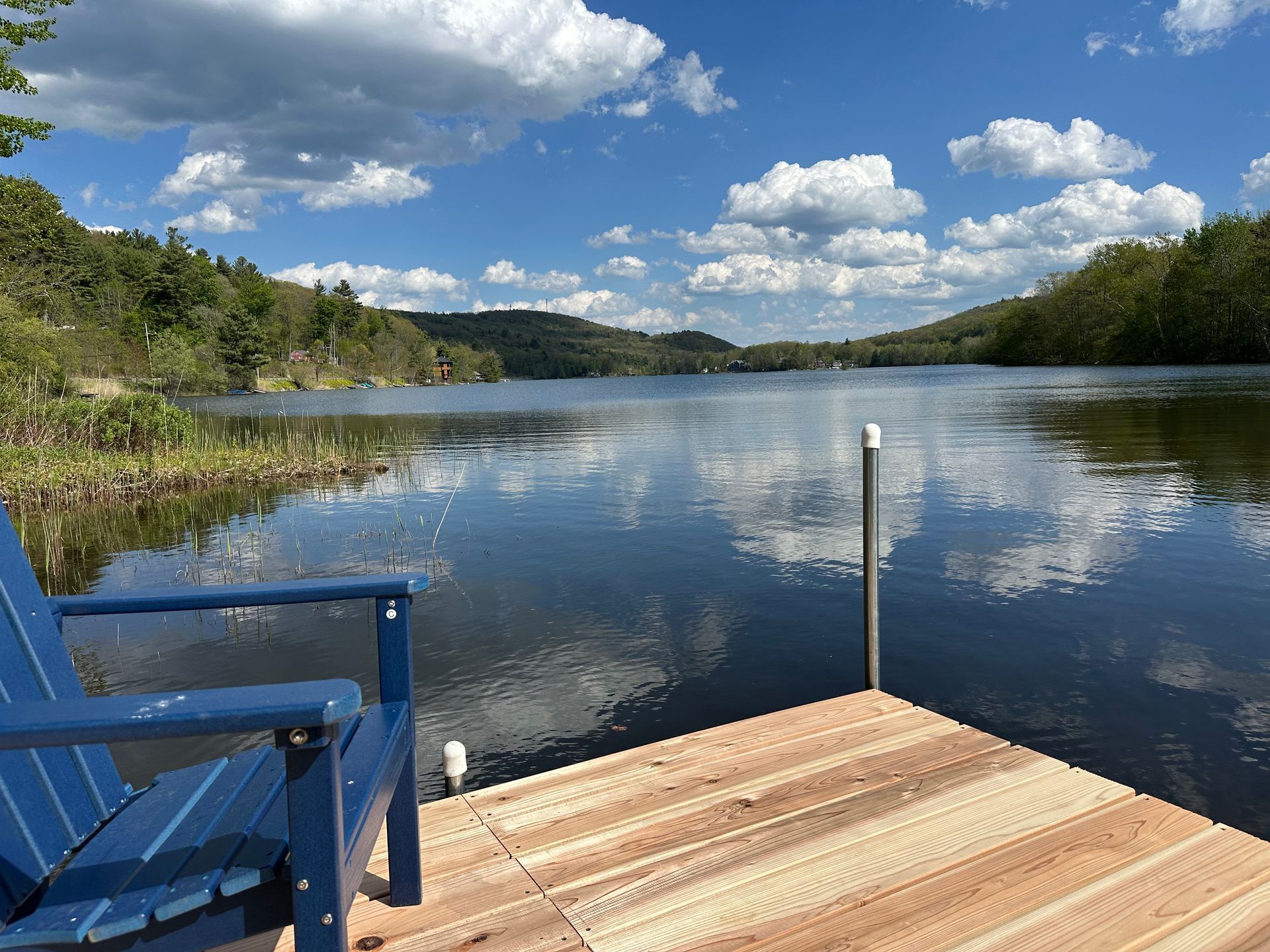 Wooden dock with blue chair overlooking a calm lake under a cloudy blue sky