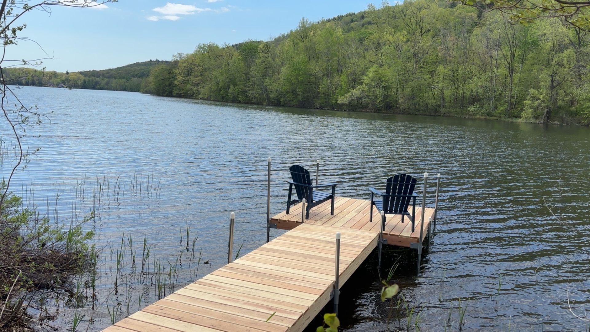 Wooden dock with two chairs extending into a calm lake, bordered by green trees under a clear sky.