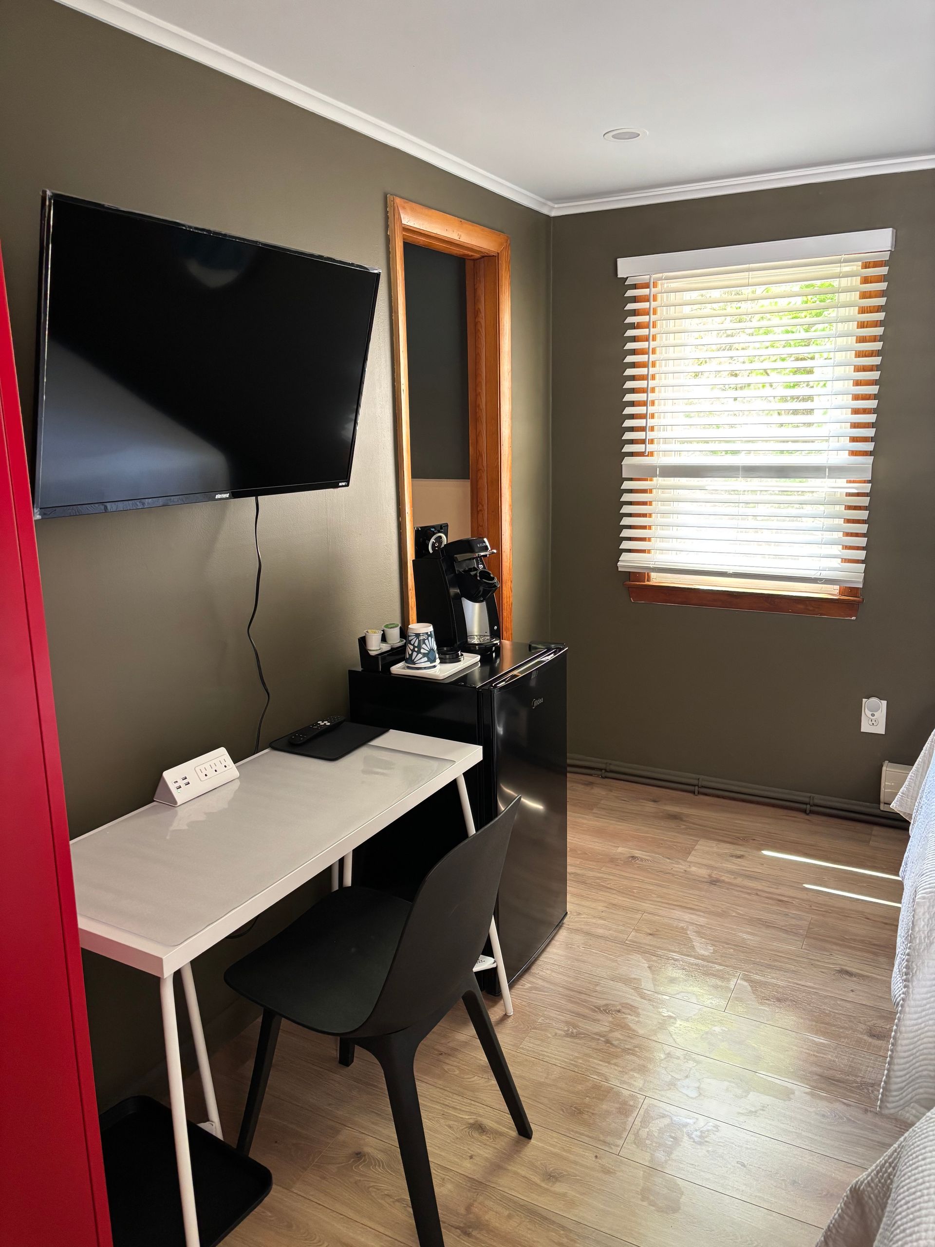 Small home office with wall-mounted TV, desk, chair, coffee machine, and window blinds.