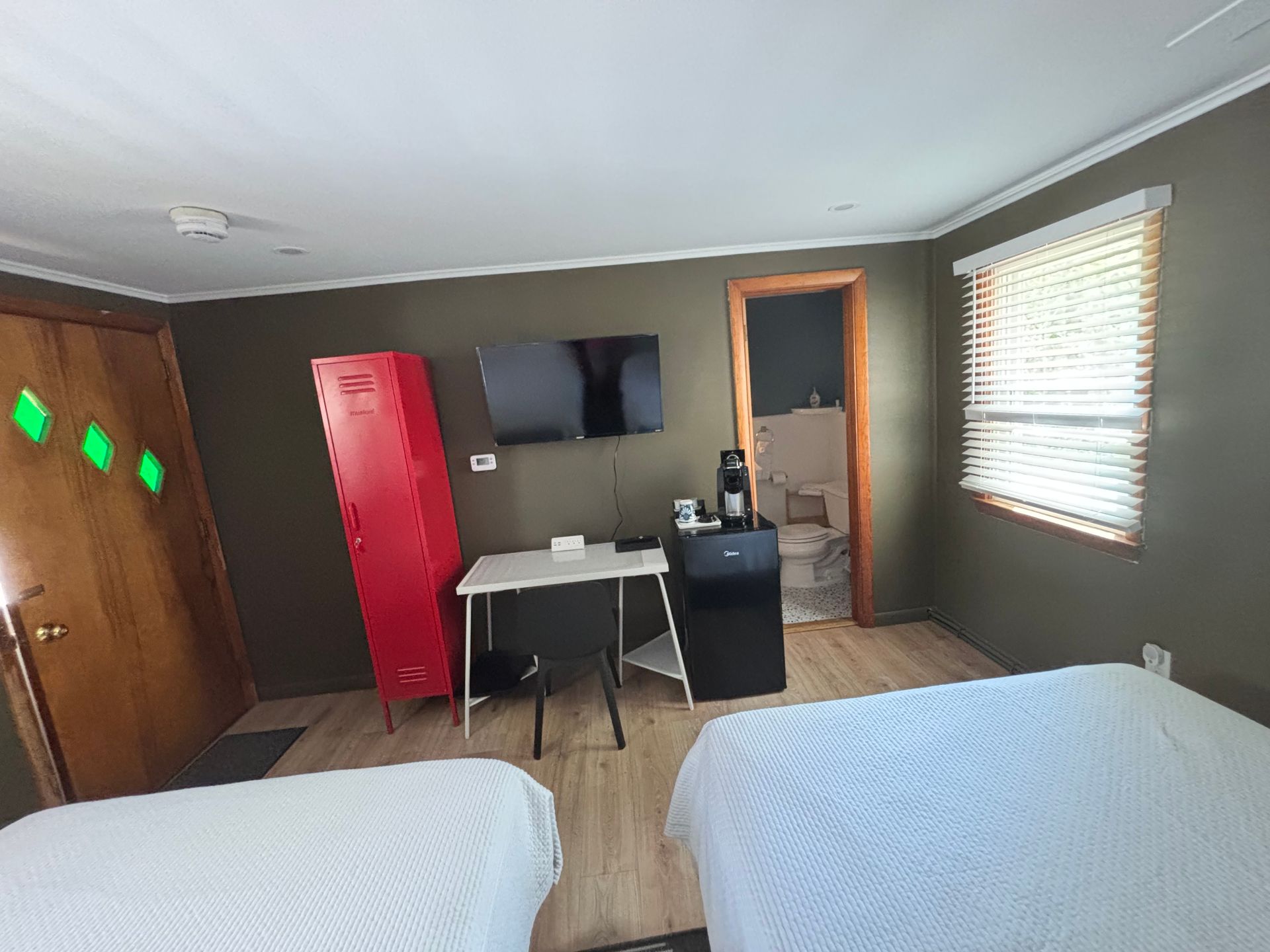 Small hotel room with two beds, red cabinet, wall TV, desk, and window blinds.