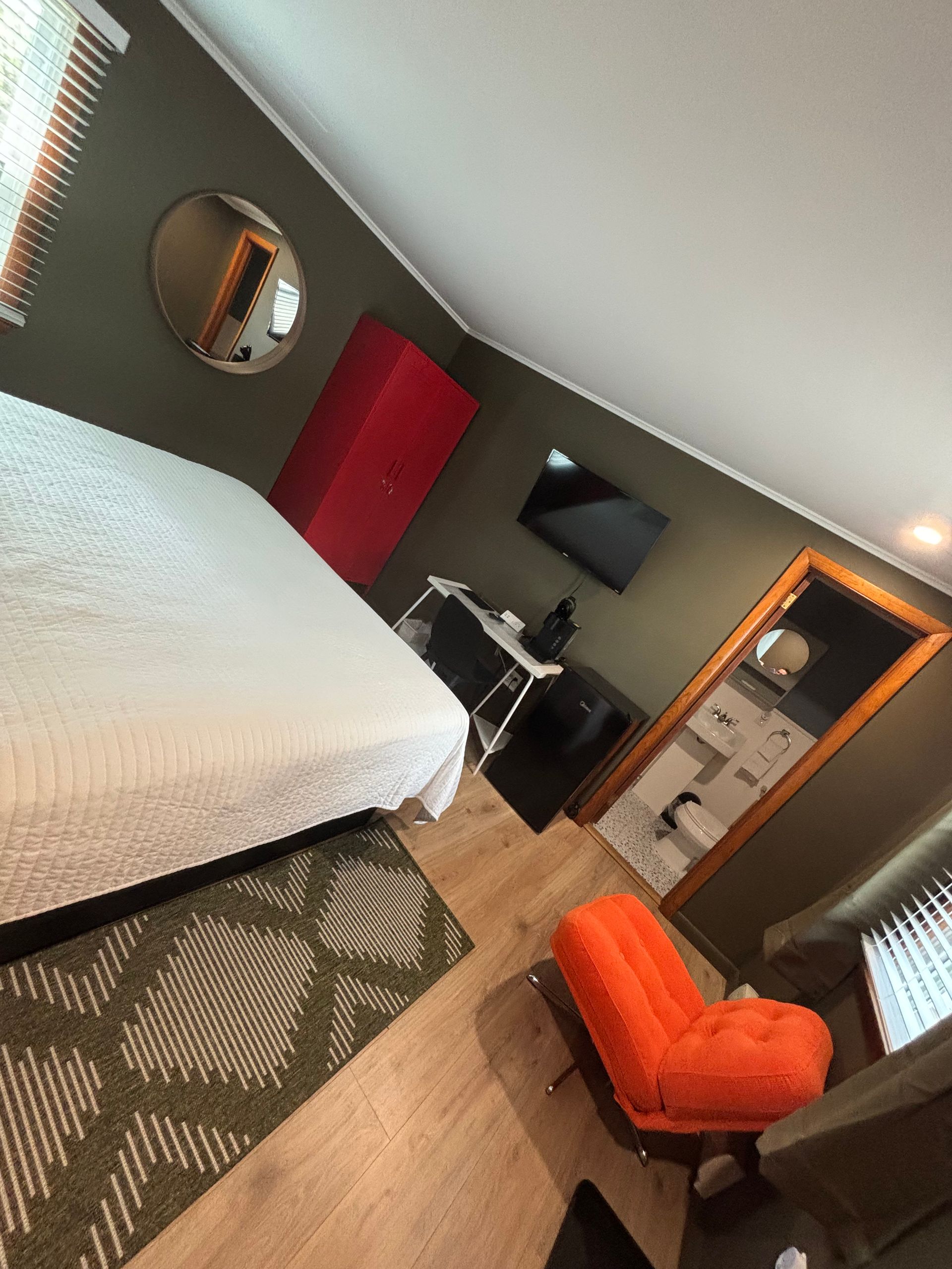 Modern hotel room with bed, orange chair, wall-mounted TV, red wardrobe, and patterned rug.