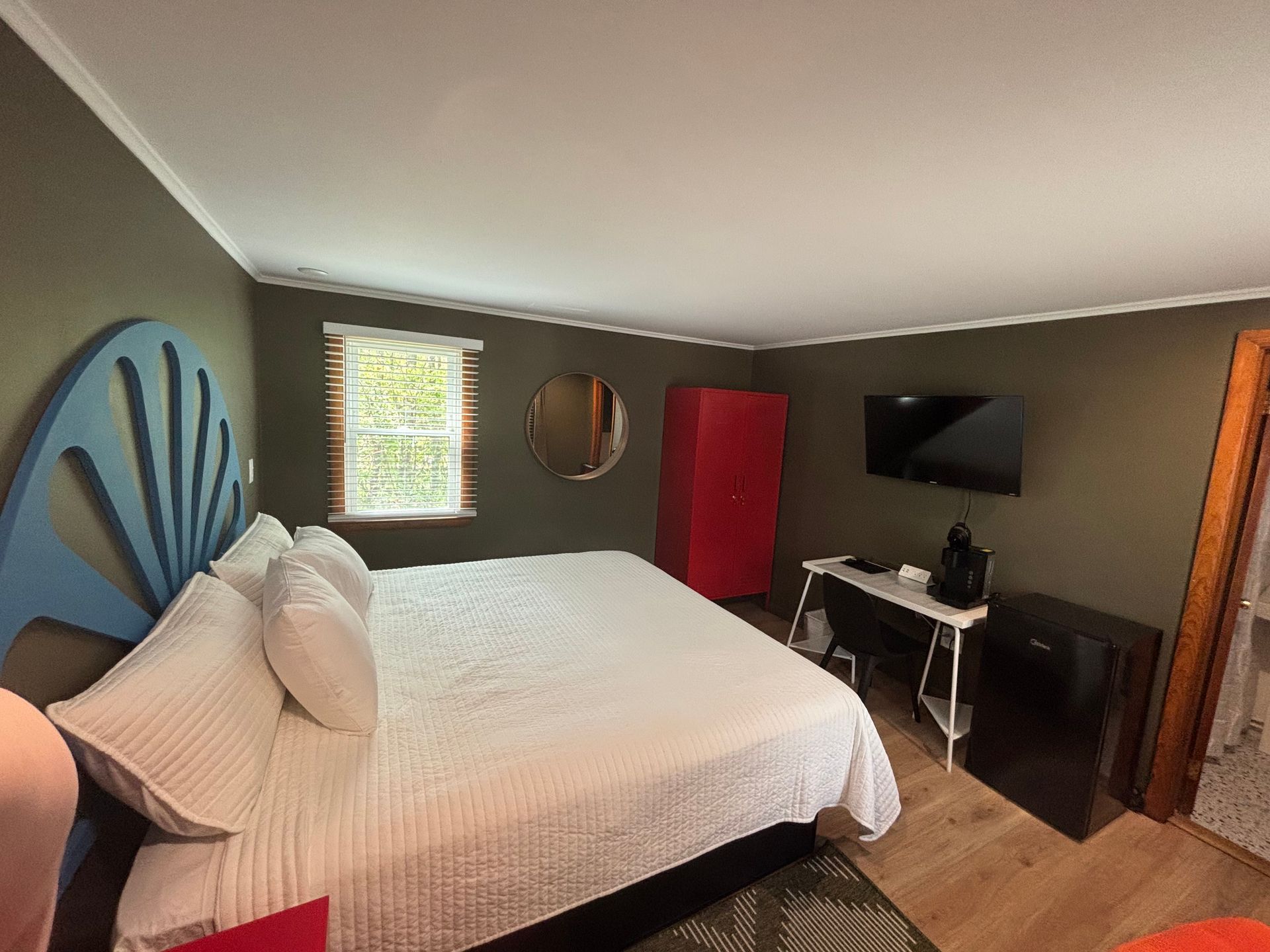 Bedroom with a bed, wall-mounted TV, small desk, mirror, and window with blinds.