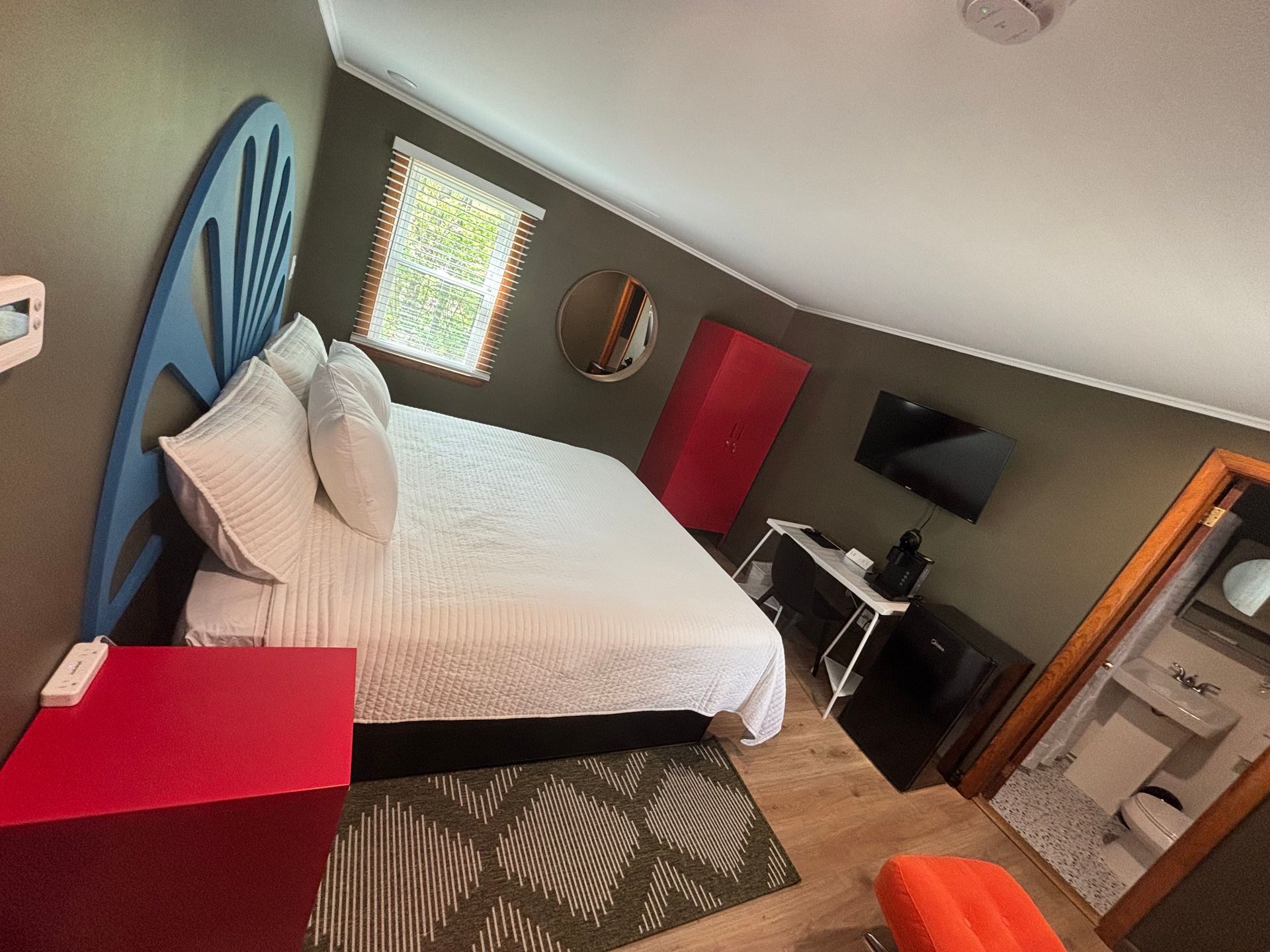 Bedroom with a blue headboard, white bedspread, red bench, desk, TV, and patterned rug.