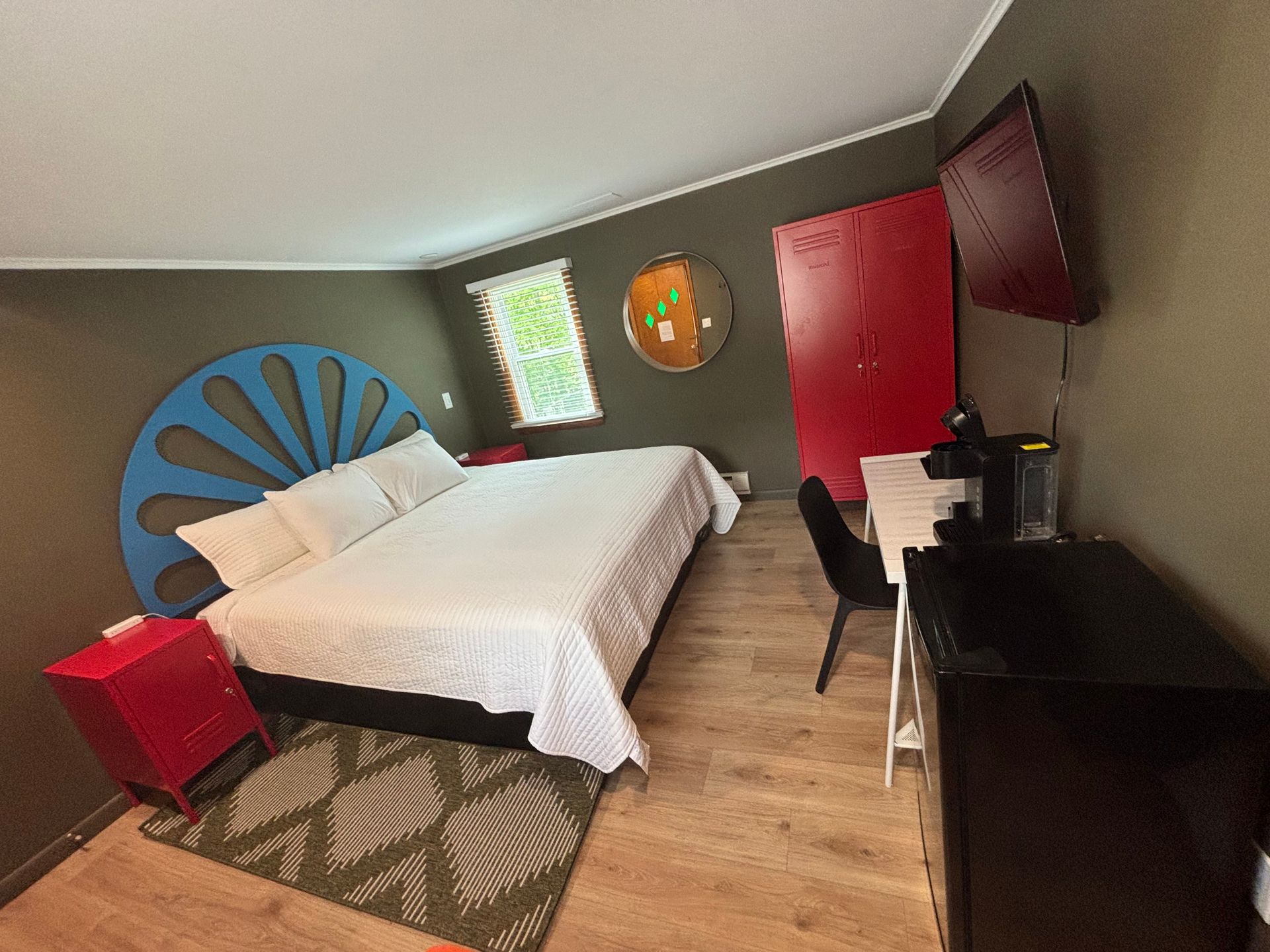 Small hotel bedroom with a white bed, blue sunburst headboard, red accents, and dark wood furniture.