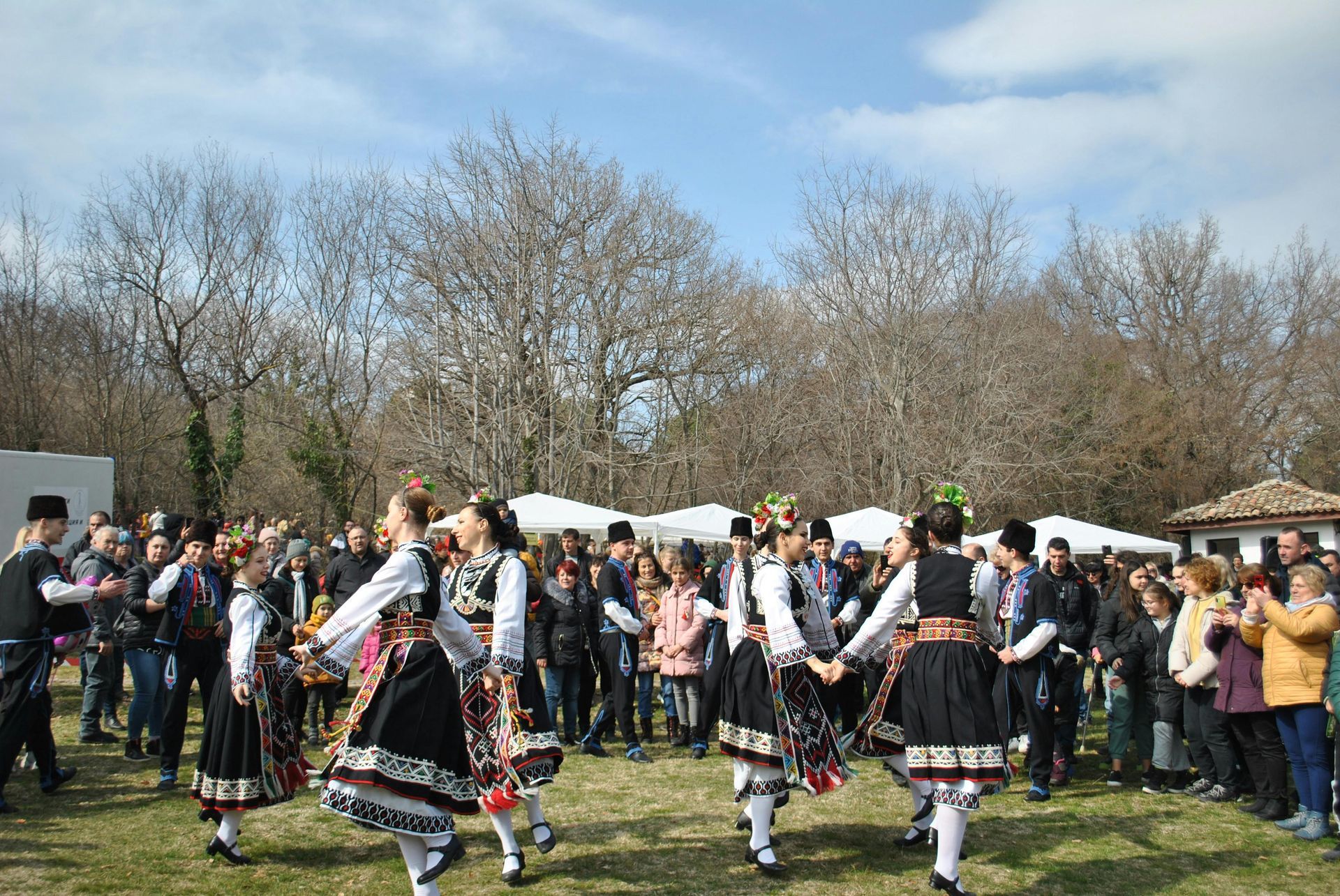 Traditional dancers in black-and-white costumes perform outdoors before a crowd in a grassy park.