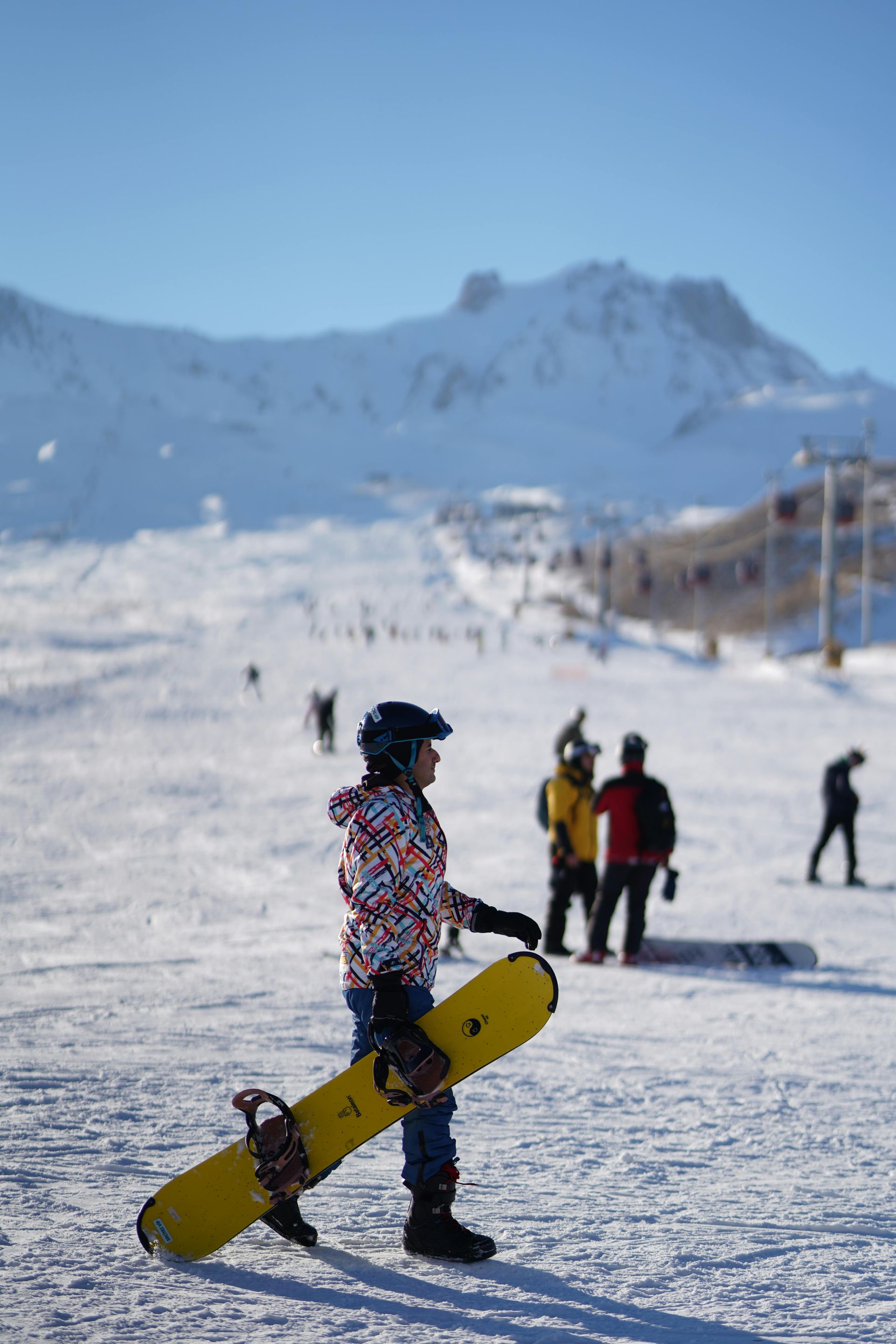 Snowboarder in colorful gear carrying a yellow board on a snowy mountain slope