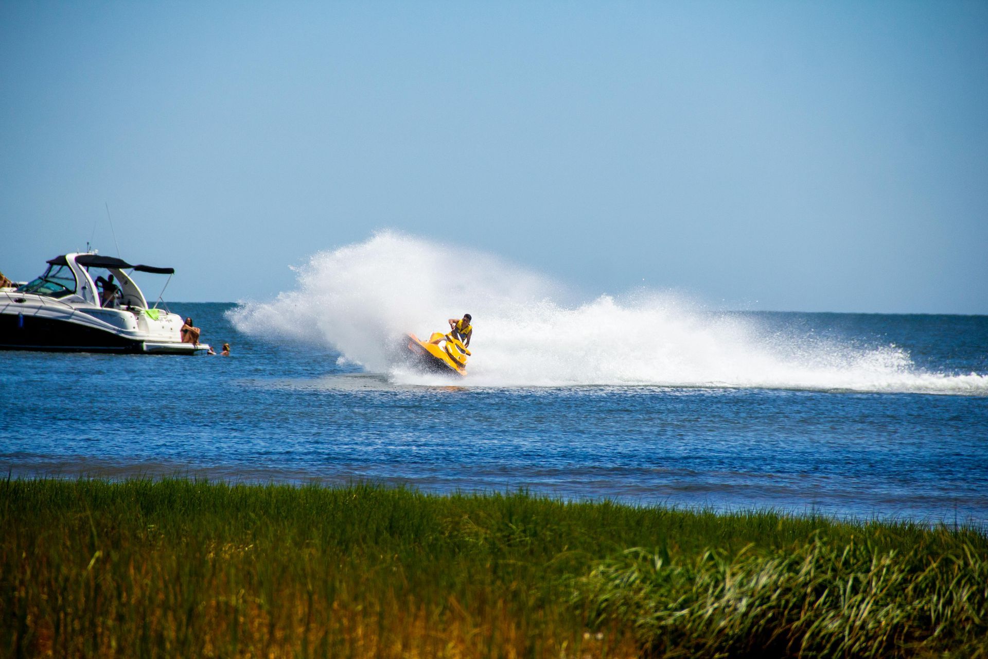Jet ski racing across blue water, spraying white foam near a yacht and grassy shore