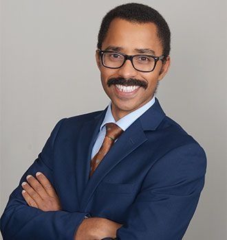 Man in blue suit, glasses, and mustache smiles with arms crossed.