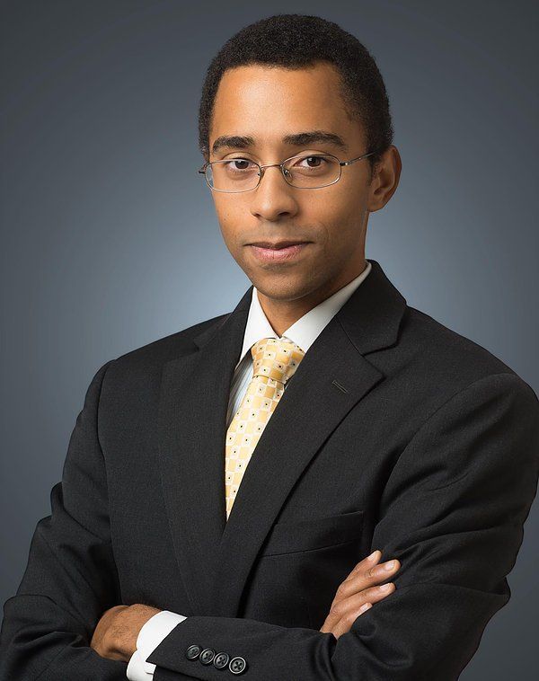 Man in blue suit, glasses, and mustache smiles with arms crossed.