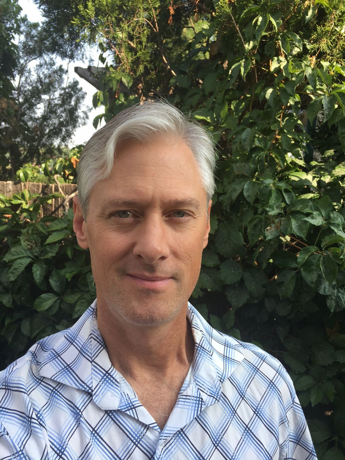 A man, Christian Jett, in a plaid shirt is standing in front of a bush.