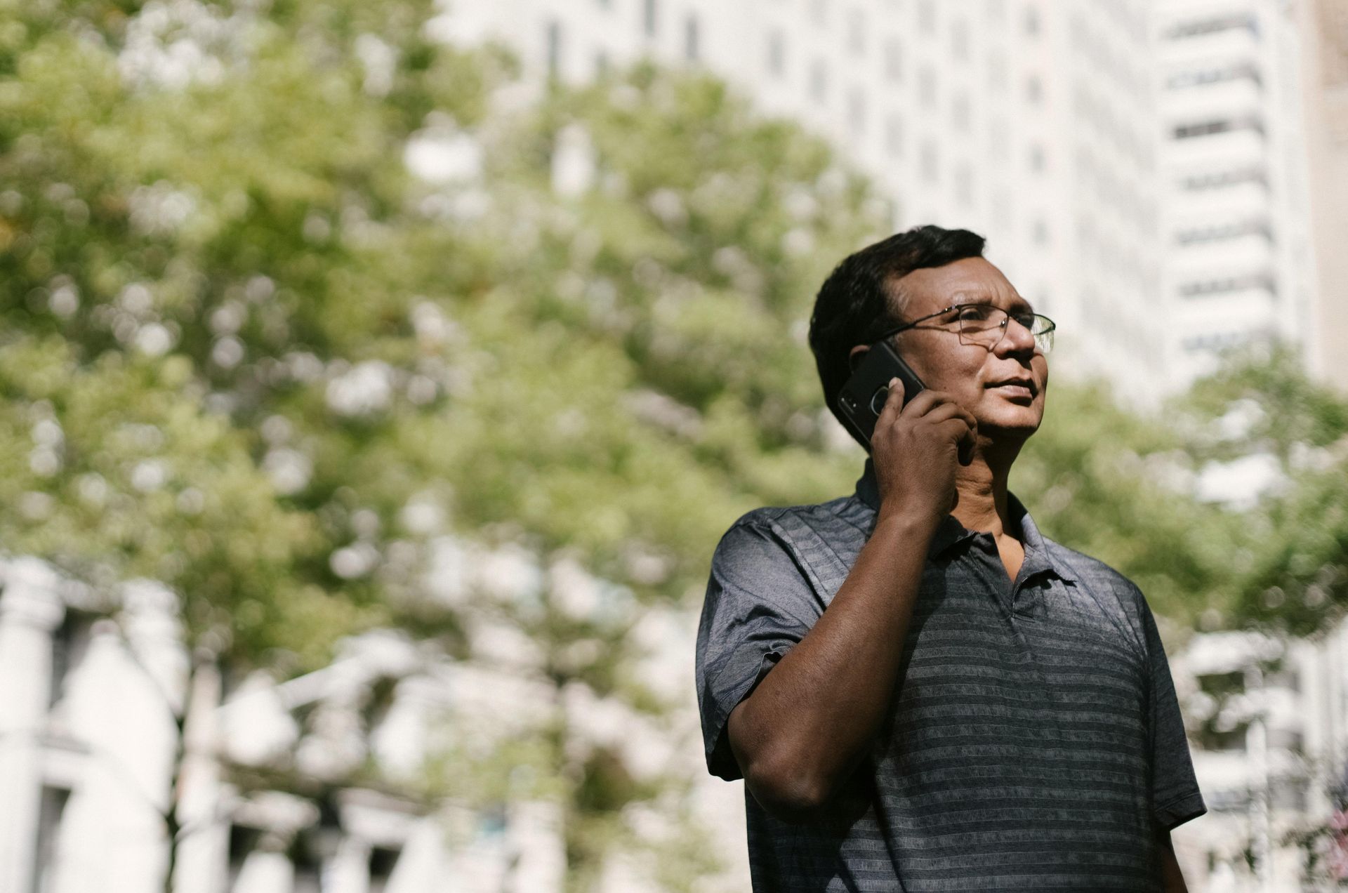 Man in glasses talking on a phone outdoors, trees and city buildings in the background.