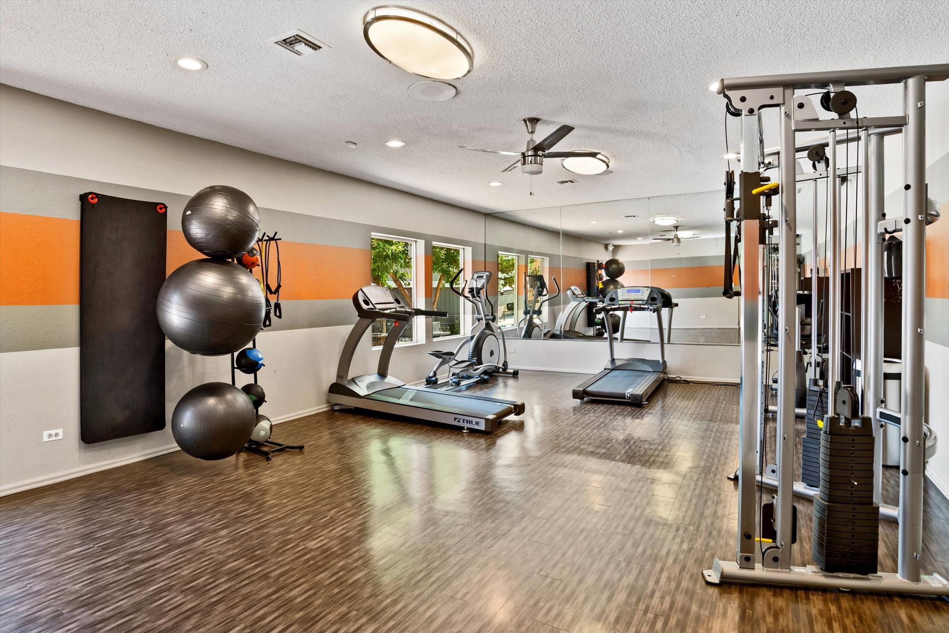 Gym interior with exercise equipment: treadmills, weights, balance balls. Orange and gray wall accents.