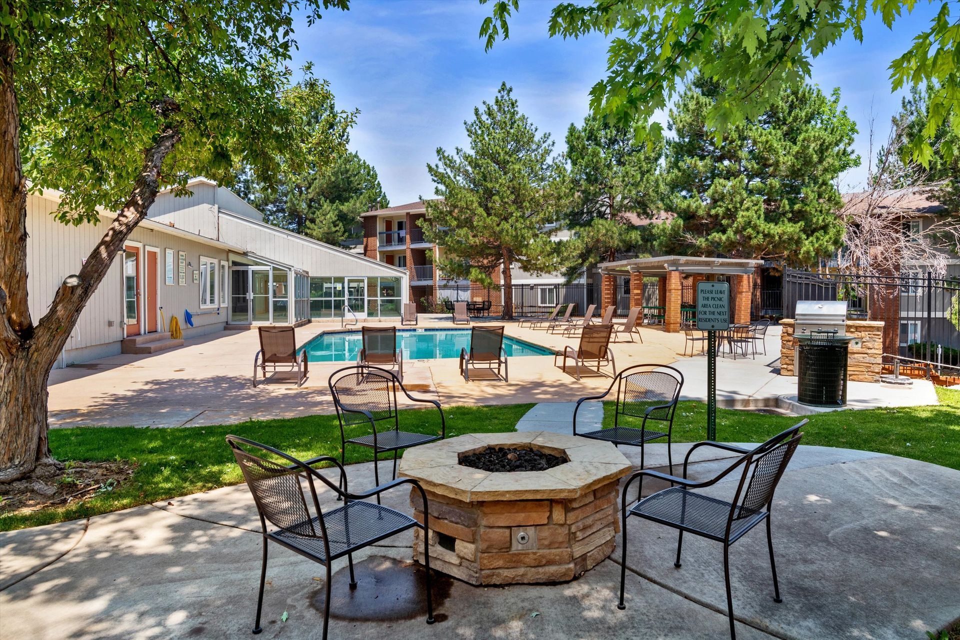 Outdoor lounge area with fire pit, pool, seating, and trees on a sunny day.