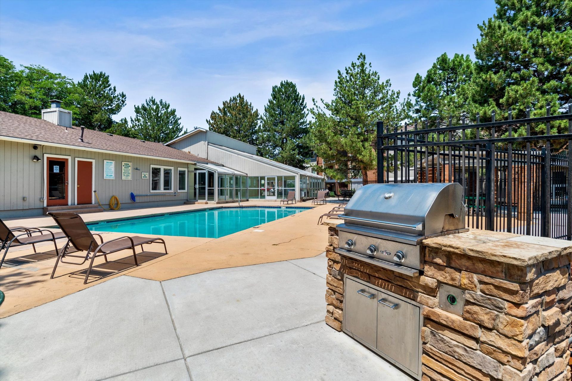 Backyard with a swimming pool, grill, and house. Tan house, blue pool, stone grill. Sunny day.
