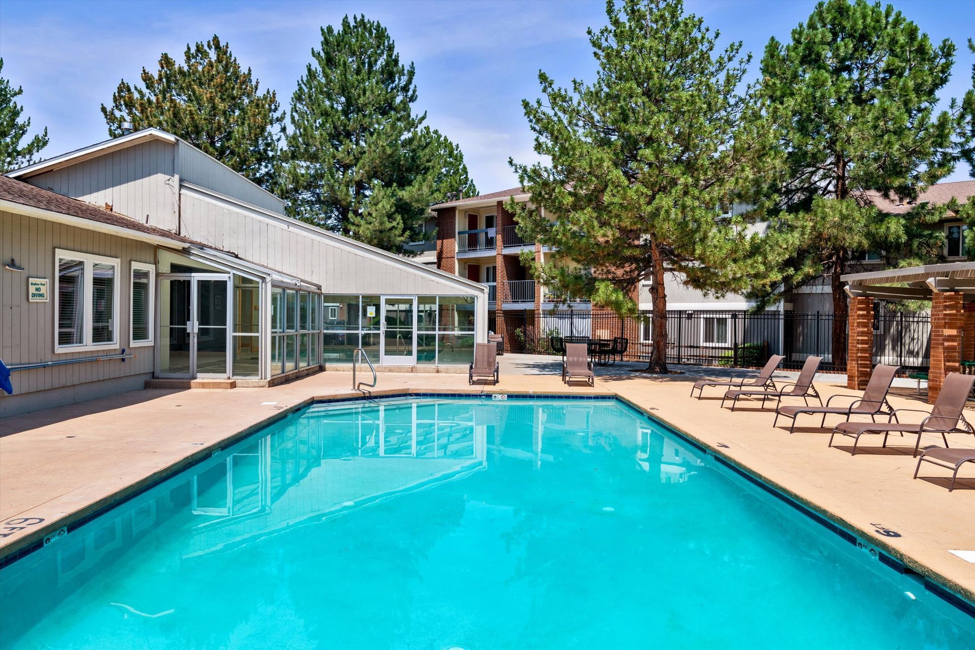 Swimming pool surrounded by lounge chairs, patio, and buildings. Sunny day with trees in the background.