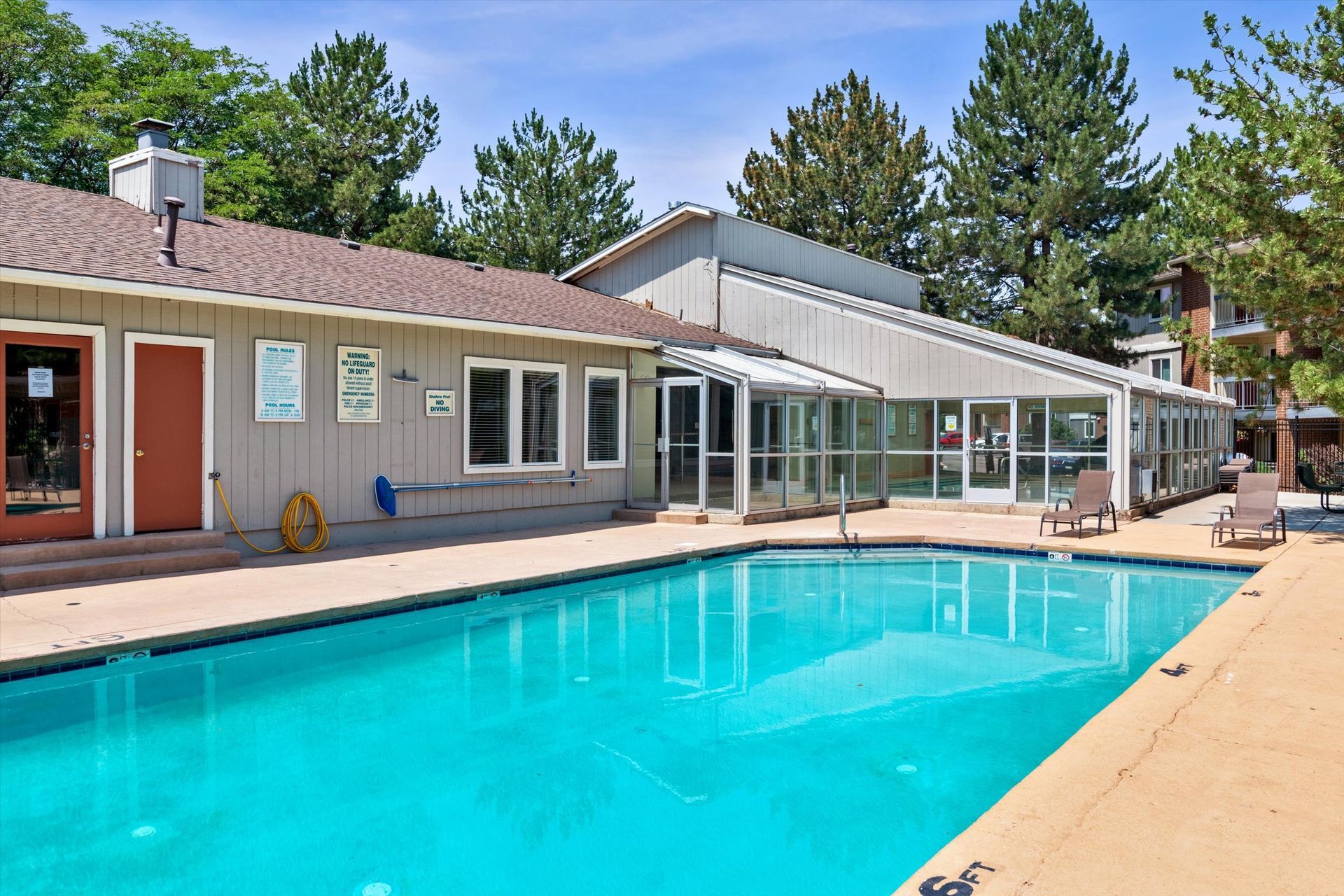 Swimming pool next to a building with glass walls, brown roof, and trees under a blue sky.