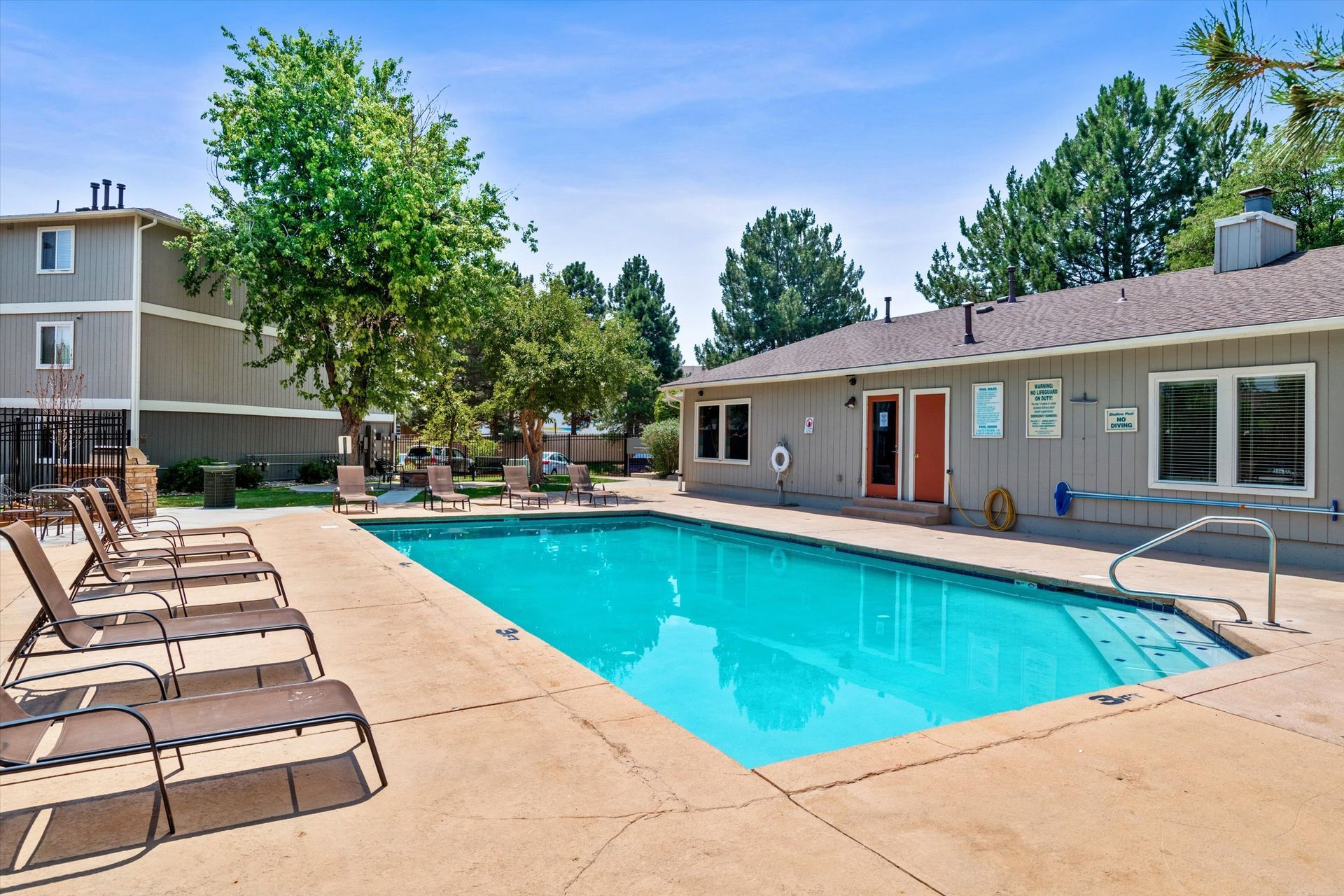 Pool with lounge chairs, trees, and buildings under a blue sky.