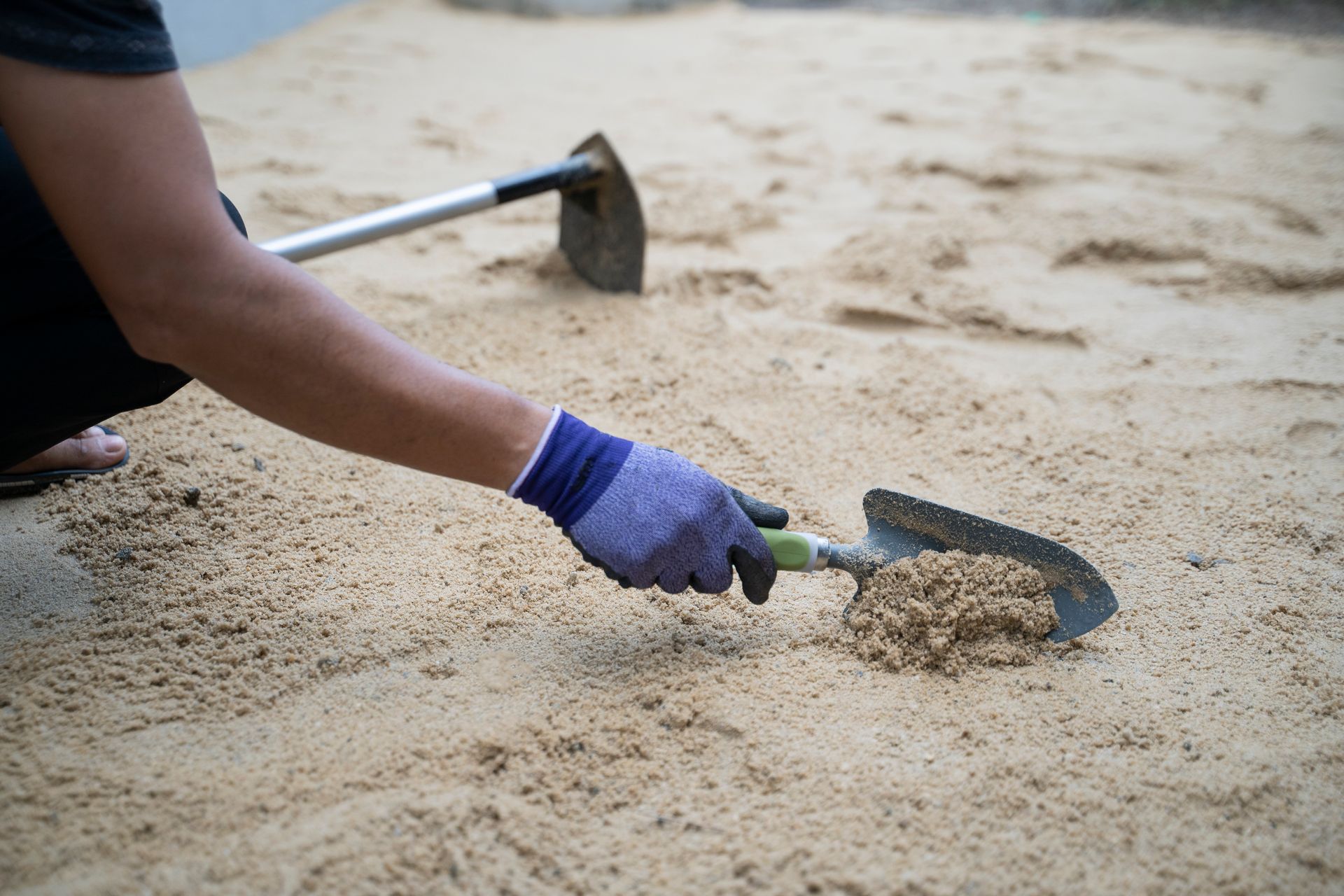 Person wearing a glove using a shovel to scoop sand, with a hoe in the background.