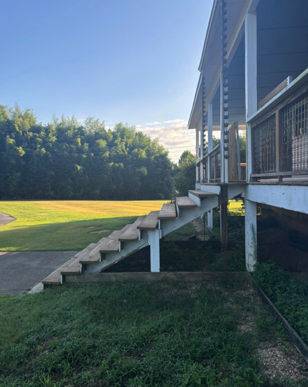Wooden stairs leading up to a porch with a railing. Green lawn and trees in the background.