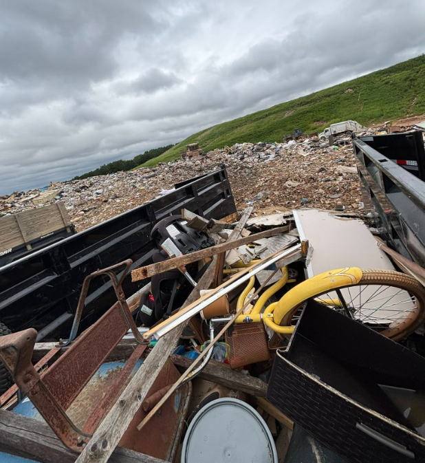 Dump truck filled with trash, against a landfill backdrop. Yellow bicycle frame visible. Overcast sky.