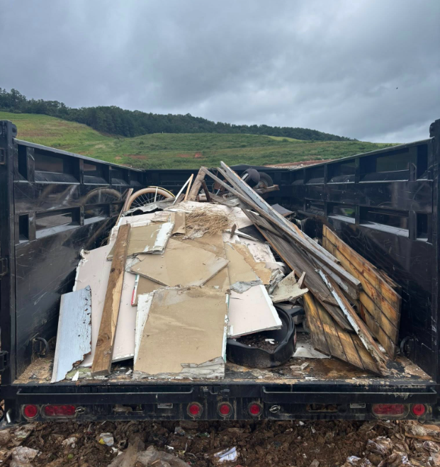 Dump truck filled with construction debris, including drywall, wood, and tires, against a hilly backdrop.