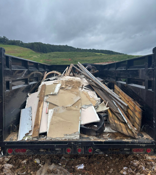 Dump truck bed filled with construction debris; cloudy sky, green hill in the background.
