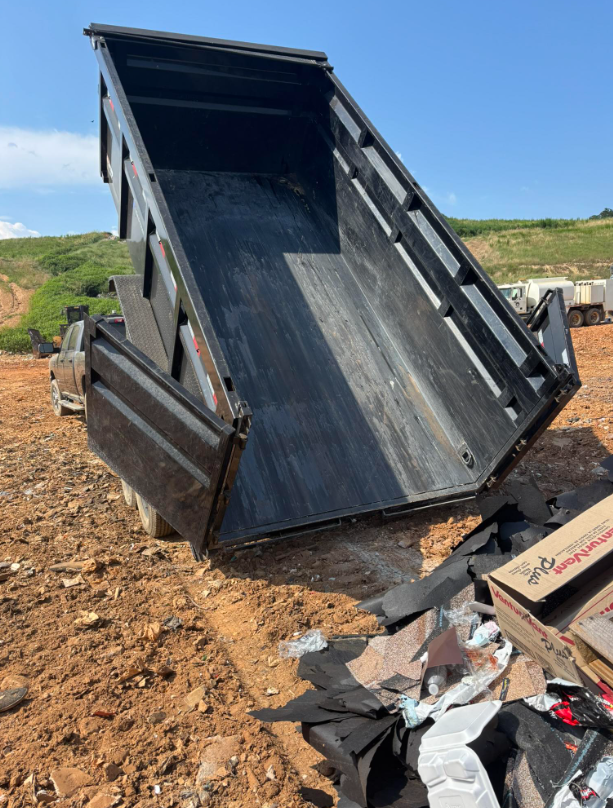Black dump truck bed tilted up, dumping debris onto dirt ground under blue sky.