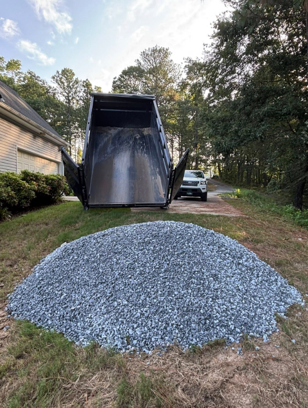 A truck bed tilted, unloading a large pile of gray gravel onto the grass.