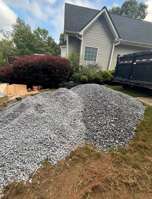 Two large piles of gray gravel in front of a house, with a red bush and a dump truck in the background.