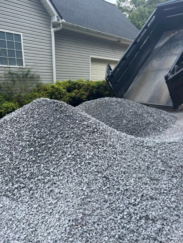 Dump truck unloading a pile of gray gravel near a house with a dark roof.