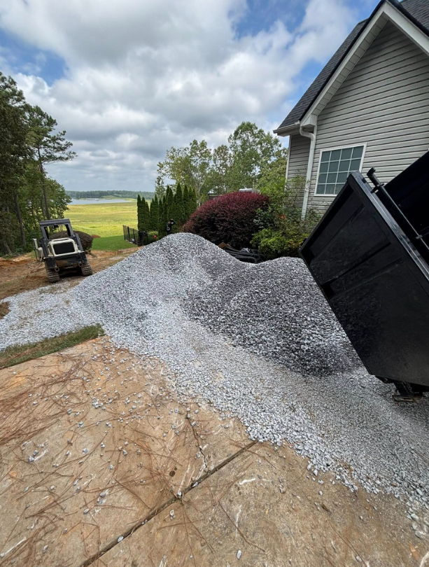 Orange dump truck unloading dark soil next to a sidewalk. Green trees in the background.
