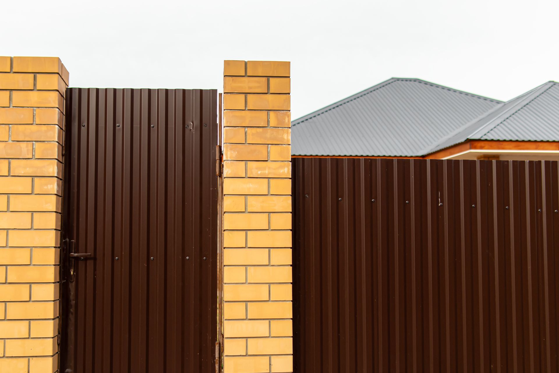 Brown metal gate and fence with brick pillars, against a gray sky, near a house with a gray roof.