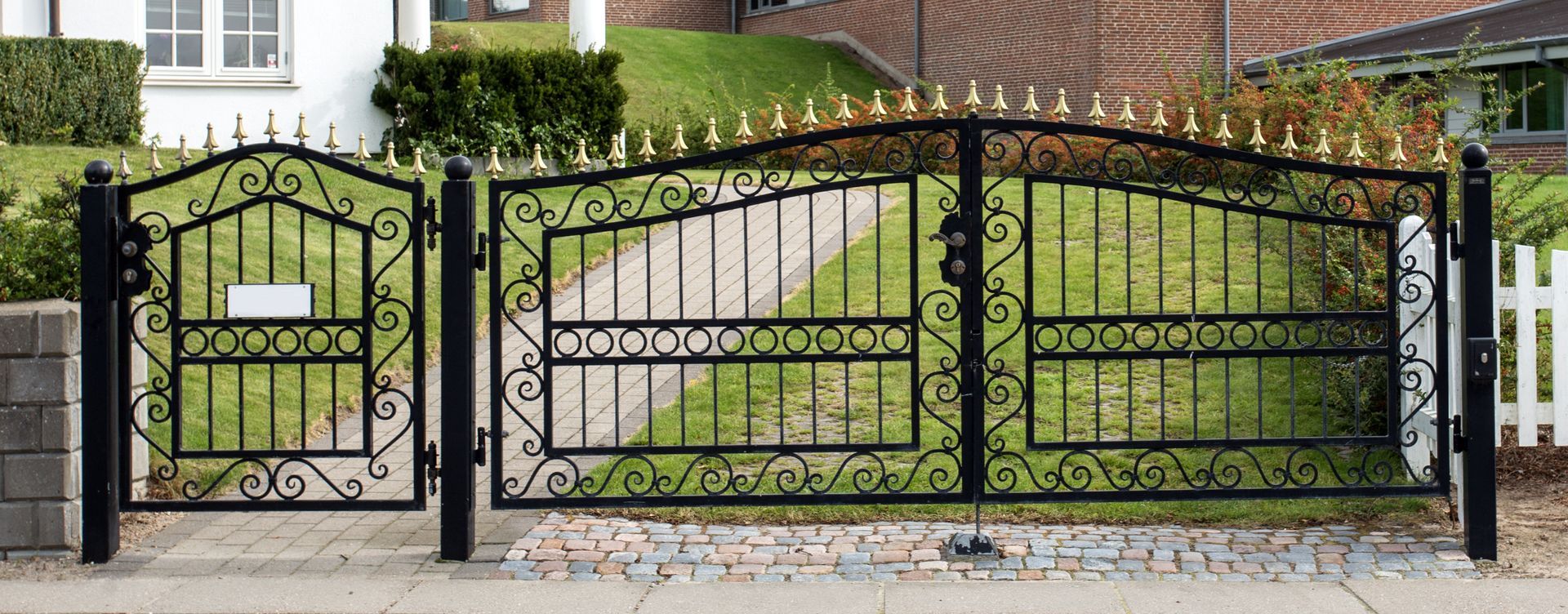 Black wrought iron gate with gold finials, mailbox, and white picket fence.
