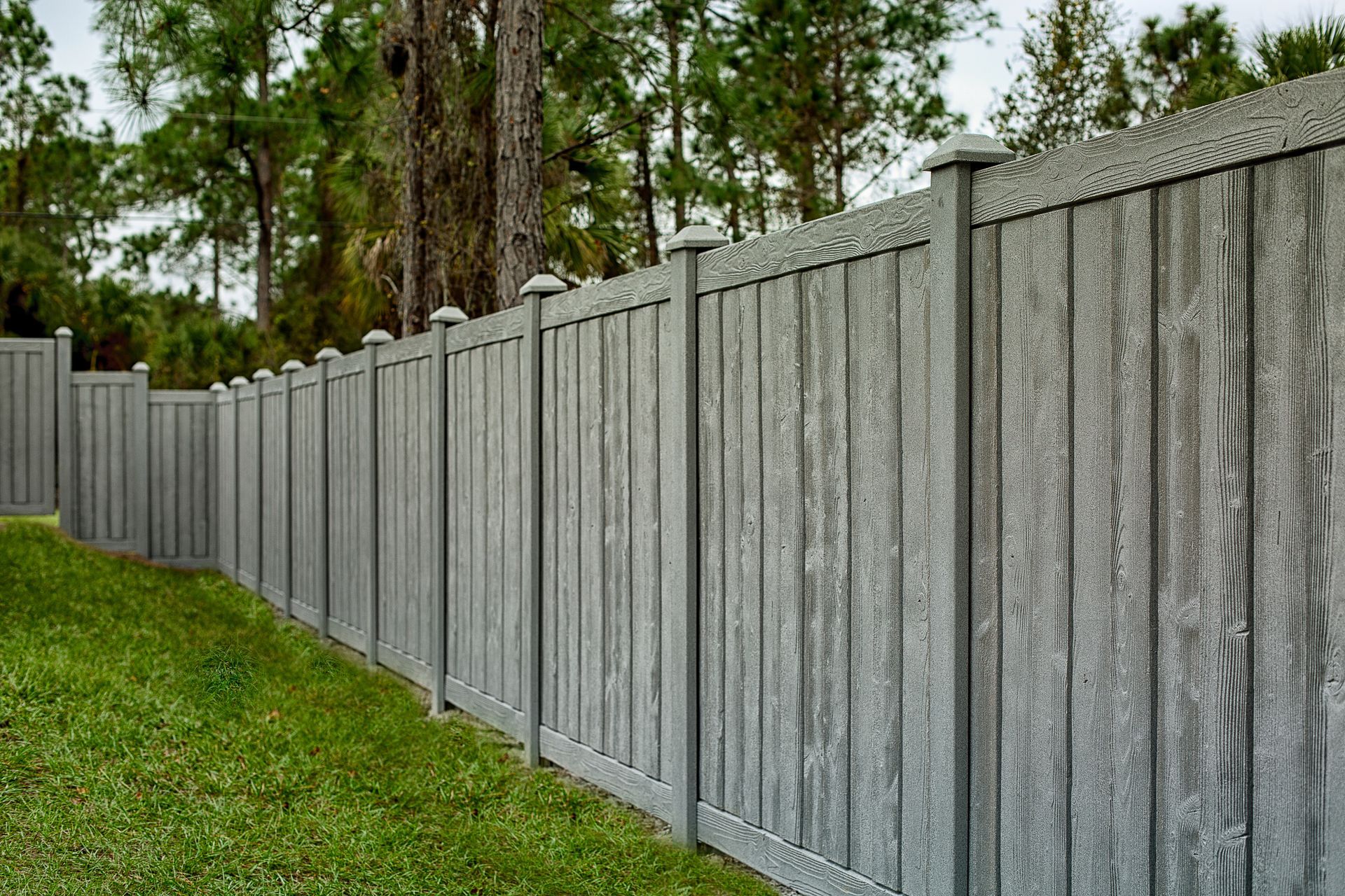 Gray composite privacy fence in a grassy yard, trees in the background.