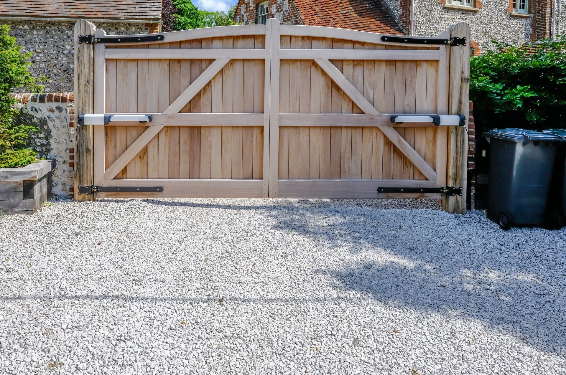 Wooden driveway gate, closed, on gravel; brick and stone house in background.