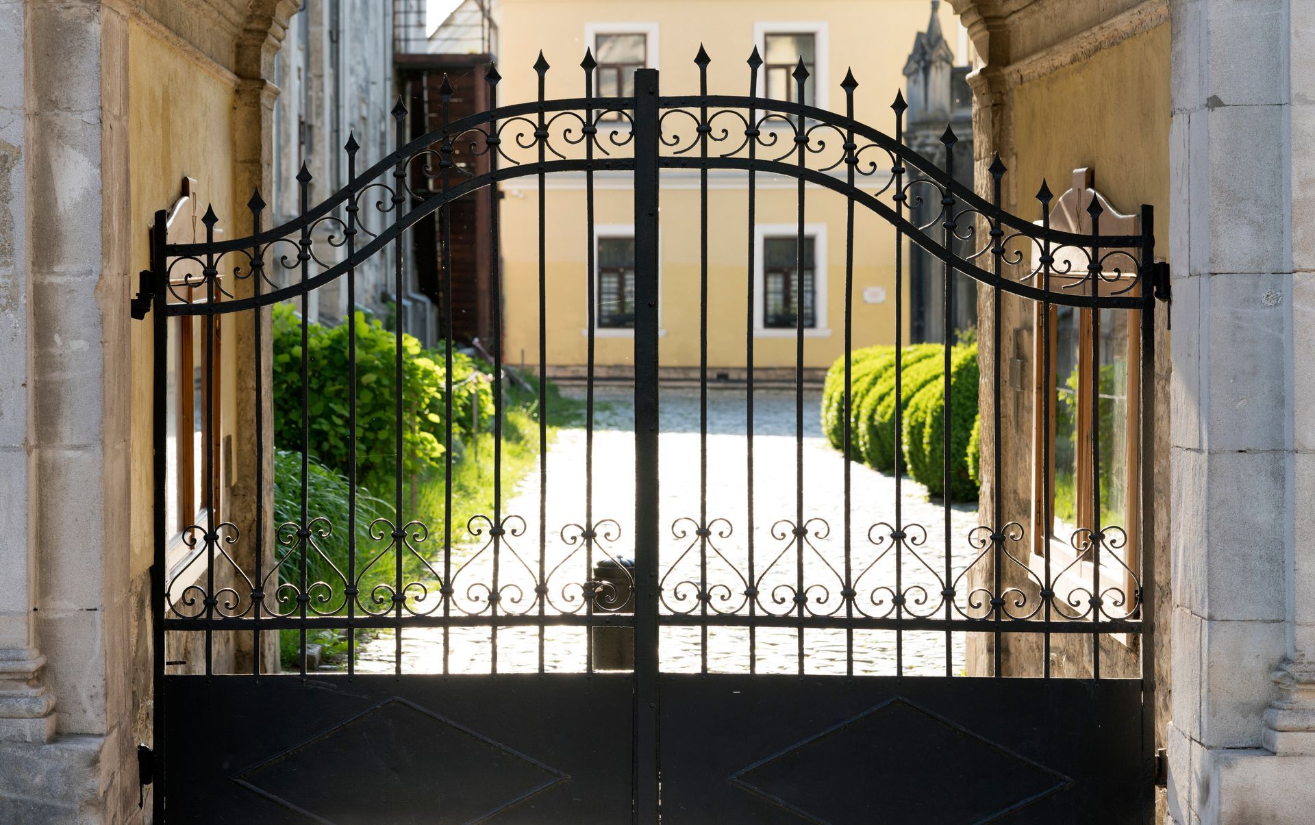 Black wrought-iron gate within stone archway, leading to a courtyard with green bushes and a light-yellow building.
