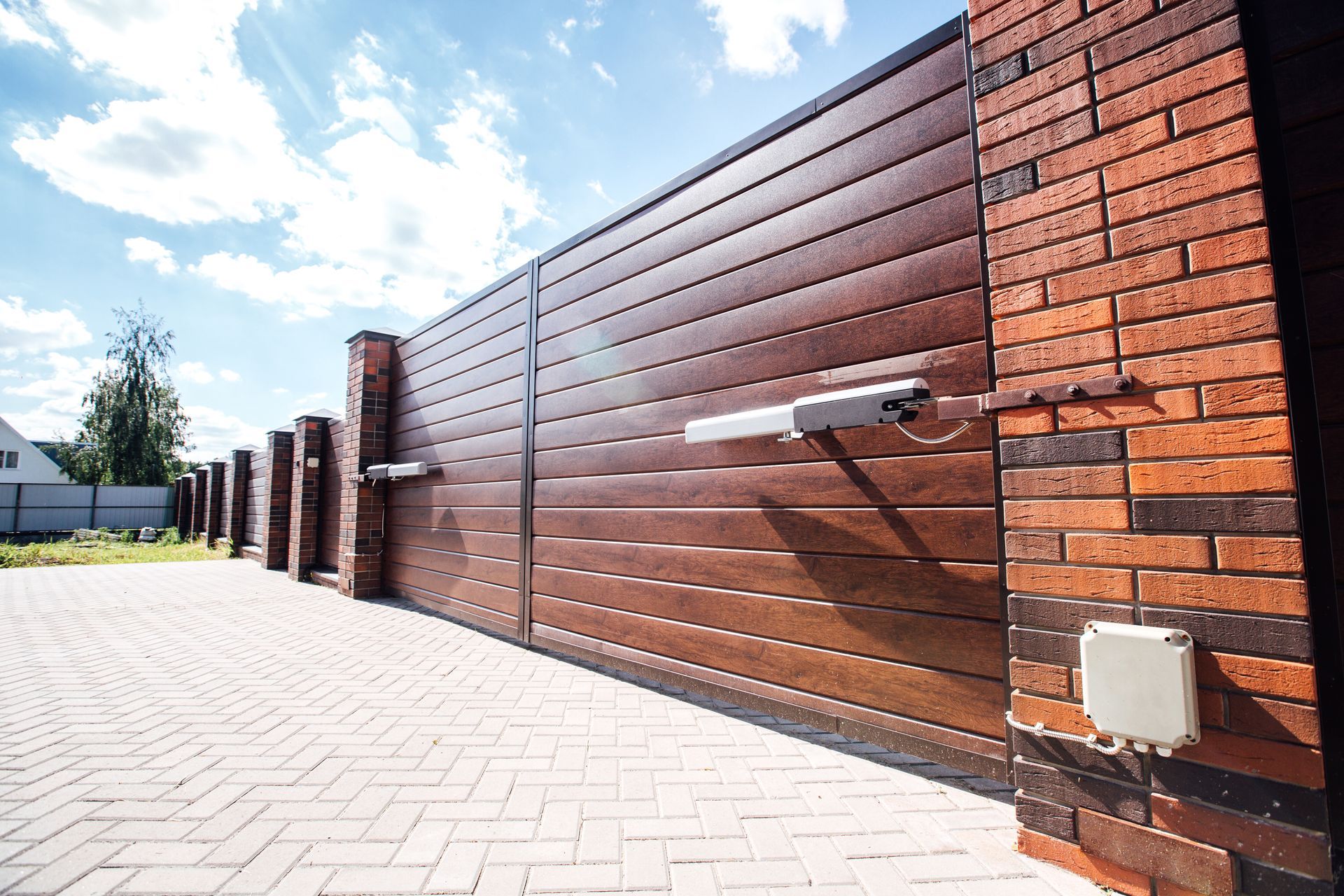 Brown sliding gate with brick pillars and control panel, driveway, and blue sky.