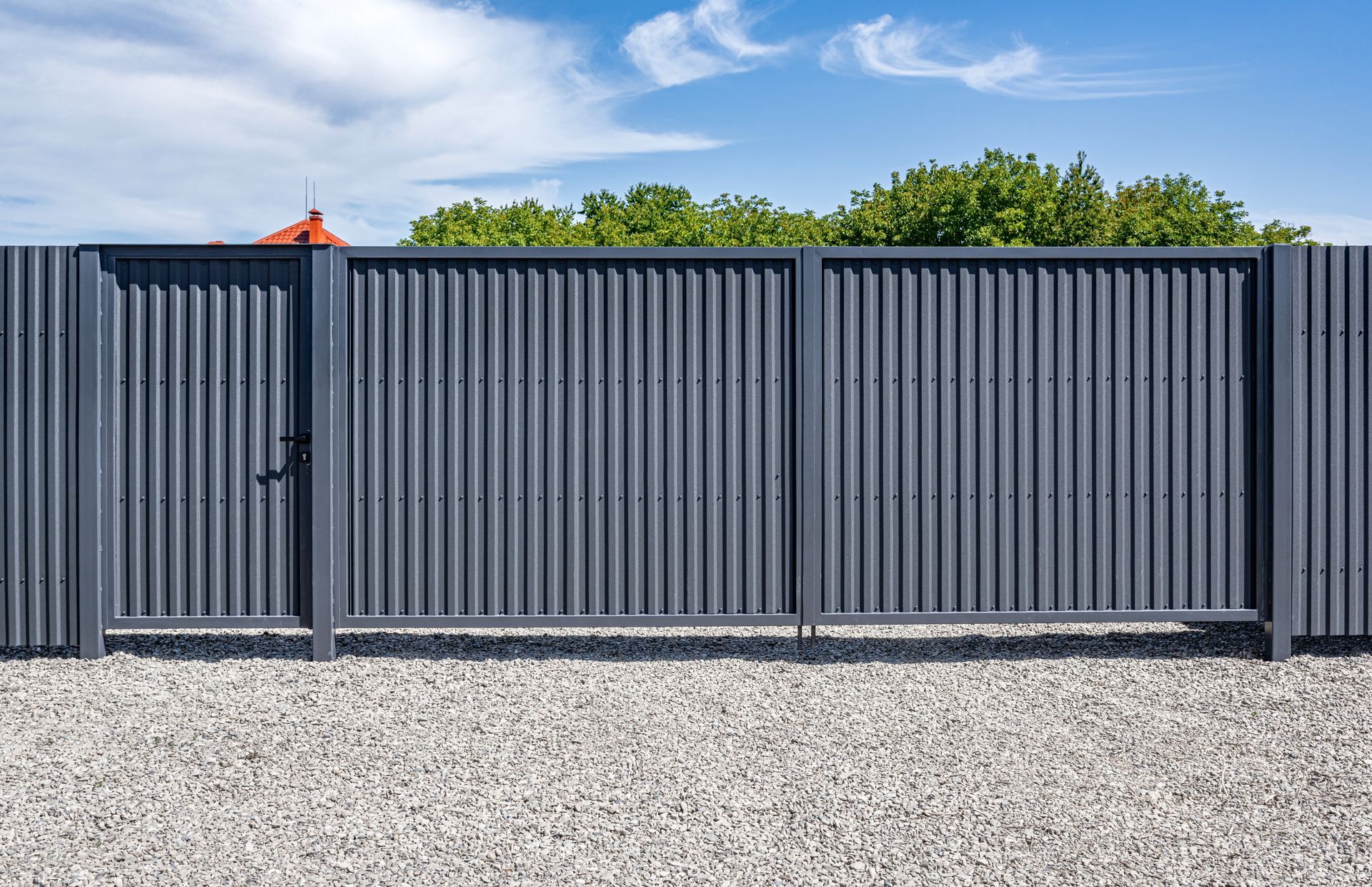 Gray corrugated metal fence and gate on gravel, with trees and sky in background.