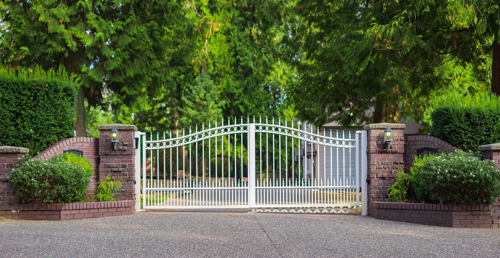 White wrought-iron gate, flanked by brick pillars with shrubbery, leading to a gravel driveway, with lush green trees in the background.