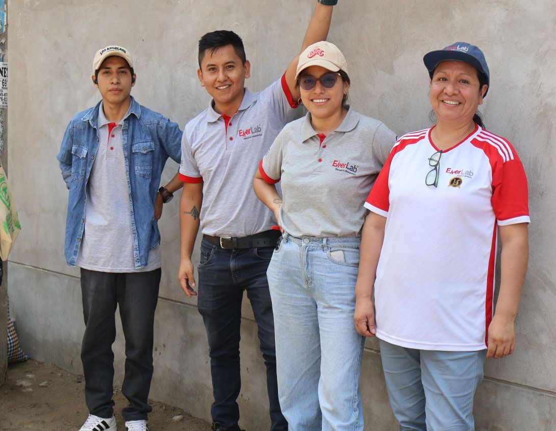 Cuatro personas están de pie frente a una pared gris; dos hombres y dos mujeres vestidas con ropa informal, sonríen.