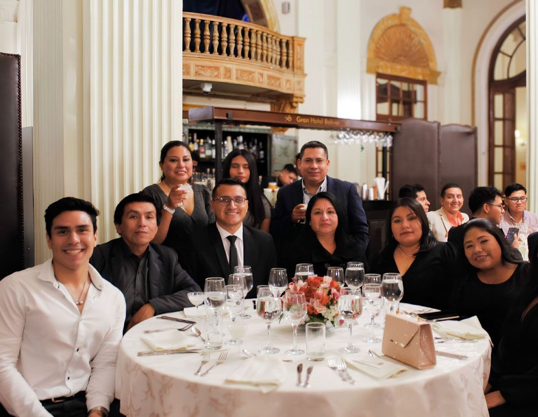 Grupo de personas en una cena formal. Rostros sonrientes alrededor de una mesa con cristalería. Salón elegante.