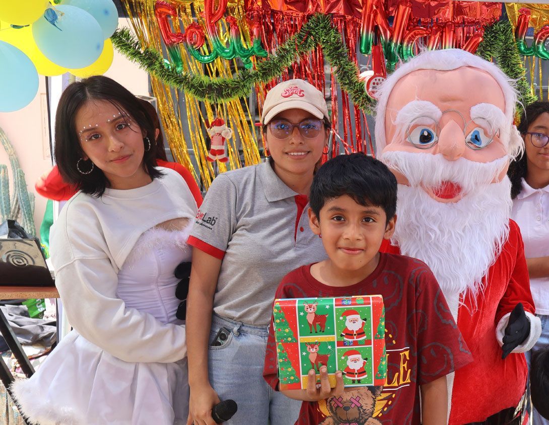 Un niño sonríe, sosteniendo un regalo envuelto con Papá Noel y dos mujeres jóvenes, en un evento navideño.