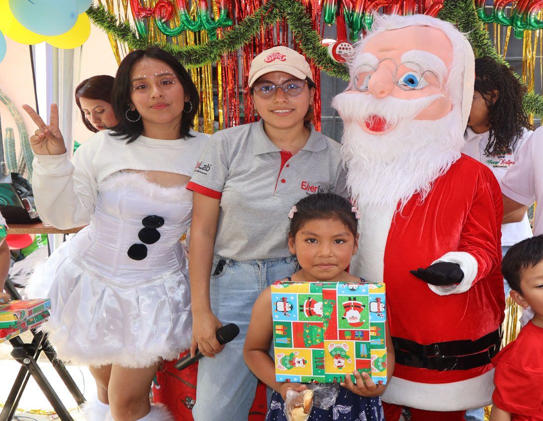 Personas con una figura de Papá Noel y un niño con un regalo. Zona decorada con globos y guirnaldas de colores.